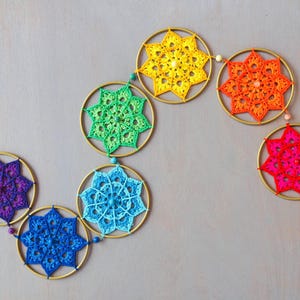 May include: A colorful, handmade crochet garland featuring seven circular designs in a rainbow of colors: purple, blue, green, yellow, orange, and pink. Each design is centered within a gold-colored metal ring and connected by a string.