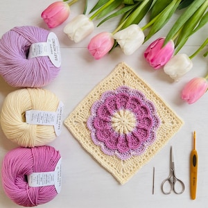 May include: A close-up of a crocheted granny square with a pink and purple flower design. The square is surrounded by yarn balls in shades of purple, pink, and yellow. The yarn balls have labels that read "Baby Merino" and "Lion Brand Yarn".