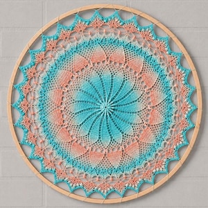 Puede incluir: Un tapiz circular con un patrón de ganchillo estilo mandala tensado en un aro de madera clara. El intrincado diseño textil muestra un esquema de color degradado que transita entre tonos turquesa, verde azulado y un suave color melocotón.