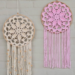 Op de afbeelding: Twee handgemaakte dromenvangers. Eén is crèmekleurig met houten kralen en de andere is roze. Beide hebben een cirkelvormig houten frame met een gehaakt middenstuk en hangende franjes.