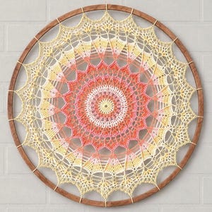 Pode incluir: Uma decoração de parede circular com um padrão detalhado de mandala em crochê num gradiente de tons amarelos, laranja e rosa. A complexa arte têxtil está esticada dentro de um aro de madeira castanho escuro, criando um design simétrico.