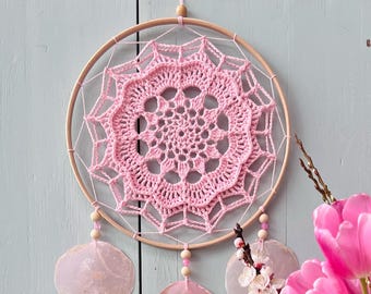 Puede incluir: Un atrapasueños circular con un tapete de ganchillo rosa claro centrado en un aro de madera. El diseño incluye hilo rosa claro, pequeñas cuentas de madera y tres discos translúcidos de color rosa pálido que cuelgan de la base.