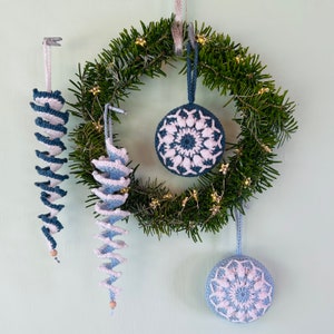 Puede incluir: Una corona de crochet azul y blanca con cuatro adornos. La corona está decorada con una guirnalda de estrellas doradas. Los adornos son un copo de nieve, una espiral y dos carámbanos.