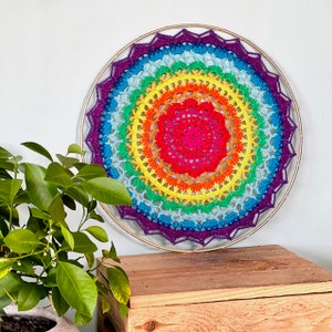 Può includere: Un arazzo da parete a mandala all'uncinetto arcobaleno con una cornice in legno. Il mandala è realizzato con una varietà di colori, tra cui rosso, arancione, giallo, verde, blu e viola.