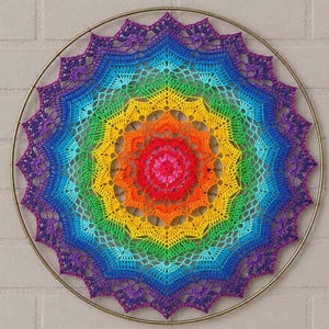 May include: A circular crochet wall hanging featuring a vibrant rainbow color gradient. The intricate mandala pattern transitions from a bright red center through orange, yellow, green, blue, and violet, all secured within a thin metallic hoop.