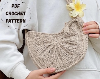 May include: A beige crescent-shaped crochet shoulder bag with a textured radial stitch pattern. A pale yellow flower-shaped hair clip is attached to the strap. The text PDF CROCHET PATTERN is printed in dark brown on the upper left side.