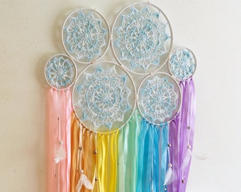 May include: A wall hanging featuring five interconnected white circular hoops containing intricate light blue and white crochet doily patterns. Long satin ribbons in a rainbow gradient of pink, orange, yellow, green, blue, and purple hang below.