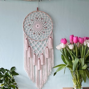 May include: A white macrame dream catcher with brown wooden beads hanging from a light blue wall. The dream catcher has a circular frame with a net design and long white fringe.