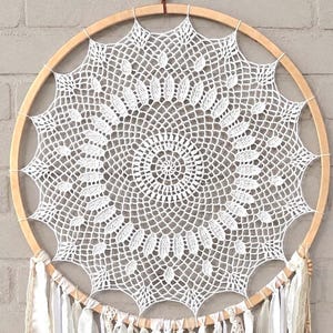 Puede incluir: Un adorno de pared circular que presenta un tapete de ganchillo blanco con un patrón geométrico complejo estirado dentro de un aro de madera clara. La parte inferior del aro está adornada con múltiples tiras de tela blanca y cintas de encaje.