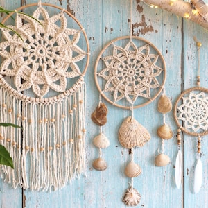 Pode incluir: Três decorações de parede circulares com mandalas de crochê cor de creme em aros de madeira. A peça da esquerda tem franjas longas com contas de madeira. A peça central apresenta conchas penduradas. A da direita inclui contas de madeira e penas.