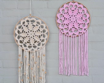 May include: Two circular wall hangings featuring intricate crochet doilies stretched over wooden hoops. The left piece is cream-colored with hanging yarn strands and wooden beads. The right piece is light pink with long, plain yarn fringe.