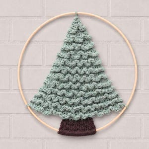 Op de afbeelding: Een wandhanger met een lichtgroene gehaakte kerstboom met een textuur van nopjessteken, bevestigd aan een ronde houten ring. De boom heeft een donkerbruine gehaakte stam aan de basis, vastgemaakt aan de onderkant van het houten frame.