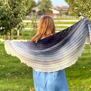 May include: A large, crescent-shaped crochet shawl featuring a gradient color transition from deep charcoal black at the top edge to a light cream white at the bottom scalloped border, showcasing intricate openwork stitches and a textured pattern.