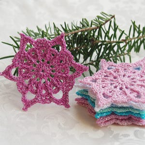 以下が含まれることがあります： ピンク、ライトブルー、ホワイトのクロシェ編み雪の結晶のセット。雪の結晶は重ねて置かれており、お祝いの華やかで繊細な外観を作り出しています。