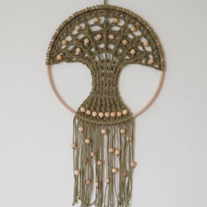 Op de afbeelding: Een ronde wandhanger met olijfgroen macramékoord geknoopt in een boomontwerp binnen een lichte houten ring. Het stuk is versierd met talloze lichtgekleurde houten kralen die zijn verwerkt in de takken en de hangende franjes aan de onderzijde.