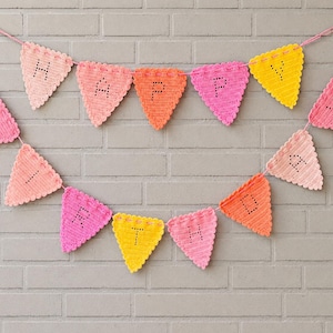 May include: A crochet bunting banner featuring triangular flags in shades of pink, orange, and yellow. Each flag displays a single black stitched letter, collectively spelling out the phrase HAPPY BIRTHDAY across two rows of hanging pennants.