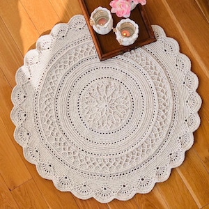 Könnte beinhalten: Ein cremefarbener, runder Häkelteppich mit einem gewellten Rand. Der Teppich weist komplizierte kreisförmige Muster und ein zentrales Blumendesign auf. Dekorative Kerzen und Blumen liegen auf einem Holztablett im Hintergrund.
