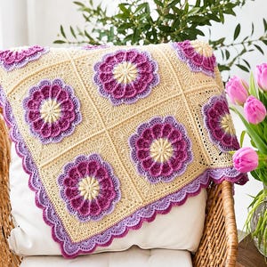 May include: A handmade crochet blanket featuring a repeating square pattern. Each square contains a central cream-coloured flower motif surrounded by vibrant magenta and light purple petals, all set against a cream-coloured background with a scalloped edge.