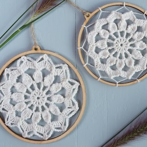 Op de afbeelding: Twee witte gehaakte dromenvangers met een bloemenpatroon. Elk is geplaatst in een houten hoepel en opgehangen met natuurlijk touw. De achtergrond is een lichtblauwe ondergrond met decoratieve planten. De diameter van elke hoepel is ongeveer 15 cm.