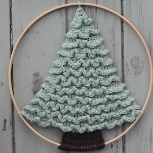 Op de afbeelding: Een gehaakte kerstboom wandhanger. De boom heeft een zachte groene kleur en is bevestigd aan een houten hoepel. De stam is donkerbruin van kleur. De boom is gemaakt met een getextureerde steek, waardoor hij een gerimpeld uiterlijk heeft.