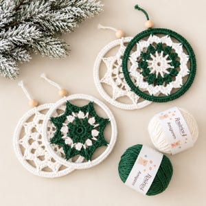 May include: Four circular crochet ornaments featuring intricate star-shaped patterns in dark green and white yarn. Each ornament is attached to a wooden bead and a hanging string. Two balls of Rosários 4 Amigarm yarn in white and green.