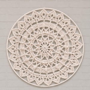 Op de afbeelding: Een ronde wandhanger met een gebroken wit gehaakt kleedje dat strak in een bijpassende gebroken witte ring is gespannen. Het ingewikkelde textielkunstwerk toont een complex, symmetrisch patroon van stralende steken en opengewerkte kantdetails.