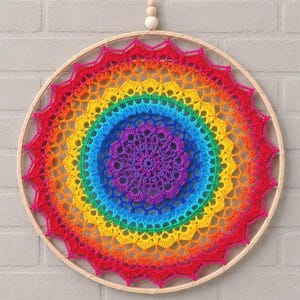 Op de afbeelding: Een ronde wandhanger met een levendig, veelkleurig gehaakt mandala gespannen in een lichtgekleurde houten ring. Het ingewikkelde garenpatroon verloopt in de kleuren van de regenboog, beginnend met paars in het midden, gevolgd door blauw, groen, geel, oranje en rood aan de geschulpte buitenrand.