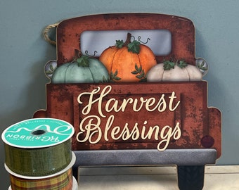 May include: A wooden truck with a red, brown, and gray paint scheme, carrying three pumpkins. The truck has the words "Harvest Blessings" written on the side. Three rolls of ribbon are in the foreground, one green, one plaid, and one with a pumpkin design.