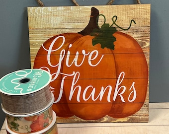 May include: A wooden sign with a rustic woodgrain background featuring a large orange pumpkin with a green stem and leaves. The sign says "Give Thanks" in white cursive lettering.