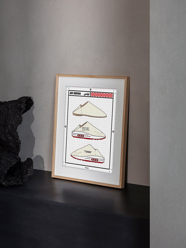 May include: Framed print featuring three illustrations of a shoe design labeled "AIR MEDINA TUNIS". The drawings are in beige and red on a grid background, with text in English and Arabic. The frame is light wood.