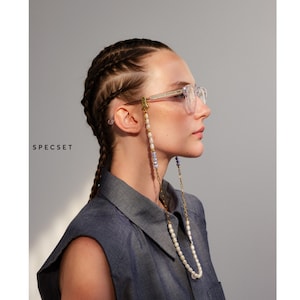 Puede incluir: Una mujer con gafas de montura transparente y una cadena de cuentas. Las gafas se sujetan con un cierre dorado. La cadena presenta perlas blancas y cuentas azules y doradas. La palabra "SPECCSET" está impresa en el lado izquierdo de la imagen.