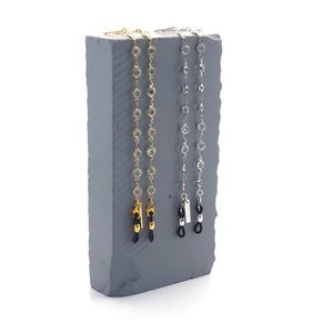 Op de afbeelding: Twee brillenkettingen met heldere, ronde kralen en metalen hardware worden getoond op een grijze, getextureerde blok. De ene ketting is goudkleurig, de andere zilverkleurig. De kettingen zijn ontworpen om brillen vast te houden.