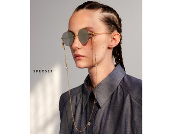 May include: Round sunglasses with a gold frame and gray lenses. A gold chain with decorative elements is attached to the glasses. The word "SPECSET" is printed in white on the left side of the image. The person is wearing a blue denim shirt.