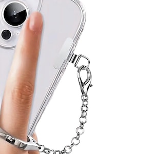 Puede incluir: Funda transparente para teléfono con cadena y anillo plateados. La funda es transparente, lo que permite ver el diseño del teléfono. La cadena y el anillo son de metal brillante.
