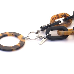 Peut inclure: Un collier en écaille de tortue noire et brune avec un grand pendentif rond. Le collier est composé de maillons épais et possède un fermoir argenté avec une petite étiquette portant l'inscription "LIVSGARD".