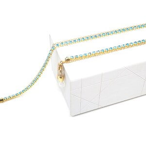 May include: A gold-toned eyeglass chain with a delicate design. The chain is adorned with small, light blue gemstones. The chain is draped over a white, geometric-patterned box, creating a contrast of colors and textures. The chain is designed to hold eyeglasses.