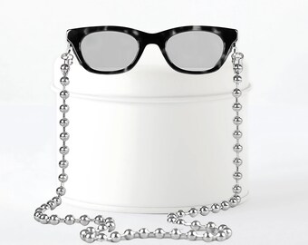 May include: Black sunglasses with a silver ball chain. The sunglasses have a classic, rectangular frame. The chain is attached to the frame and hangs down. The sunglasses are resting on a white surface.