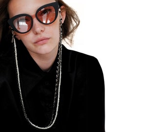 May include: Black cat-eye sunglasses with pink-tinted lenses are paired with two eyeglass chains. One chain is a delicate strand of white pearls, and the other is a silver chain. The person is wearing a black shirt.