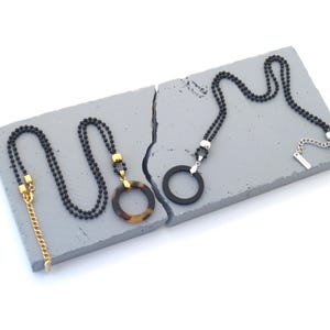 Puede incluir: Tres collares con cadenas de cuentas negras y diferentes colgantes. Uno tiene una cadena dorada y un acento cuadrado dorado, otro un colgante de carey y el tercero un colgante de anillo negro. Sobre una superficie gris.