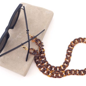 Puede incluir: Gafas de sol negras con una cadena de carey apoyada sobre una piedra gris claro. Las gafas tienen una montura cuadrada y lentes oscuras. La cadena está hecha de eslabones grandes y ovalados en tonos marrones y ámbar.