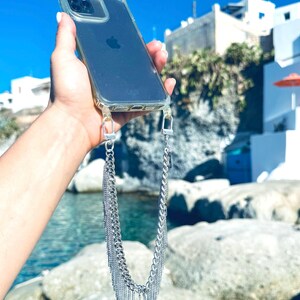 A clear phone case with a silver chain phone strap. The phone case is on a person's hand, and the chain is hanging down in front of a blue body of water and rocks.