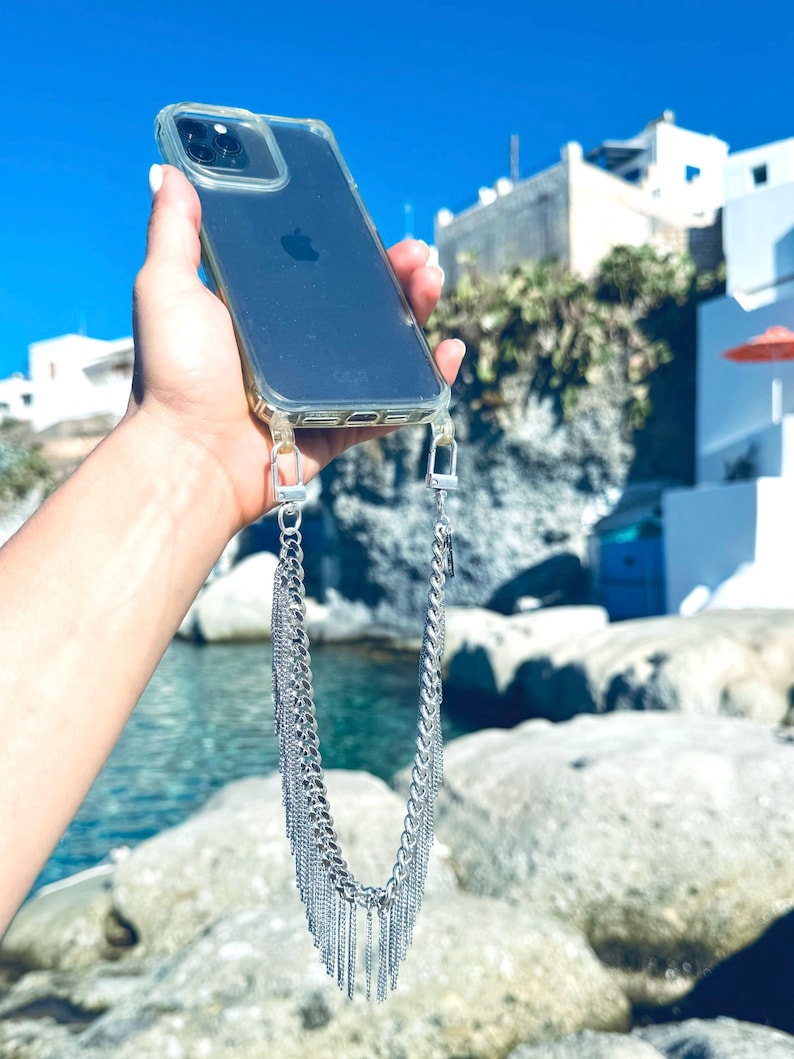 A clear phone case with a silver chain phone strap. The phone case is on a person's hand, and the chain is hanging down in front of a blue body of water and rocks.