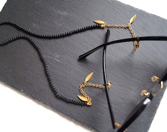 Könnte beinhalten: Eine Brillenketten aus schwarzen Perlen mit goldfarbenen Metallgliedern und kleinen blattförmigen Anhängern in Gold. Die Kette ist an den schwarzen Bügeln einer Brille befestigt, wobei die Perlen eine lange Schlaufe bilden und goldene Kettenenden.