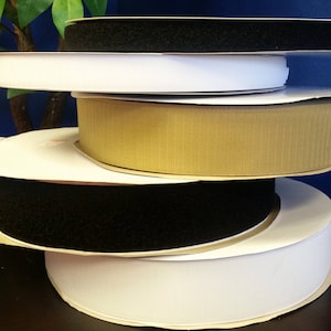 May include: A stack of five rolls of hook and loop tape in various colors, including black, white, and beige. The rolls are stacked on a dark brown surface.