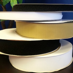 Może przedstawiać: Stosik pięciu rolek taśmy rzepowej w różnych kolorach, w tym czarnym, białym i beżowym. Rolki są ułożone na ciemnobrązowej powierzchni.