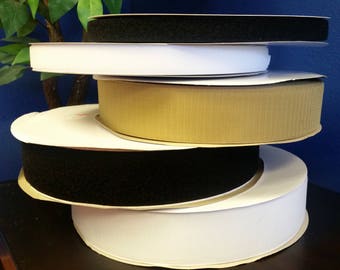 May include: A stack of five rolls of hook and loop tape in various colors, including black, white, and beige. The rolls are stacked on a dark brown surface.
