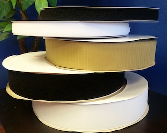 May include: A stack of five rolls of hook and loop tape in various colors, including black, white, and beige. The rolls are stacked on a dark brown surface.
