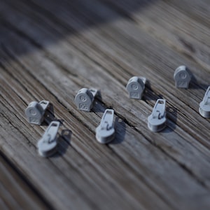 Puede incluir: Una colección de pequeños tiradores de cremallera de color gris claro dispersos sobre una superficie de madera envejecida. Los tiradores de cremallera tienen forma redondeada con un lazo y un pequeño número. La superficie de madera tiene un aspecto natural y rústico.