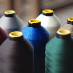 Puede incluir: Primer plano de siete carretes de hilo de varios colores, incluyendo rojo, gris, azul, negro, blanco, verde y marrón. Los carretes están dispuestos en semicírculo sobre una superficie gris.