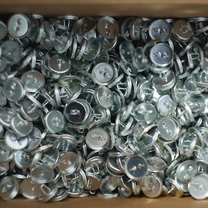May include: A close-up shot of a box filled with numerous silver metal buttons. Each button has two holes and a loop on the back for sewing. The buttons are round and have a slightly domed surface.